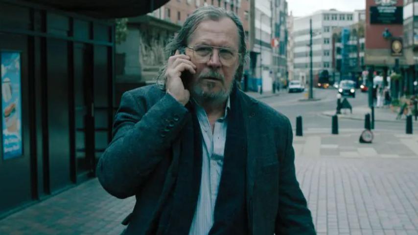 Actor Gary Oldman playing character Jackson Lamb, he is in a blue shirt, cardigan and grey overcoat while holding a phone to his ear walking down a London high street