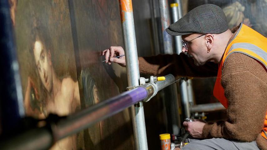 A man in a high-vis orange vest and cap sits on scaffolding, carefully working on the restoration of a large classical painting. 