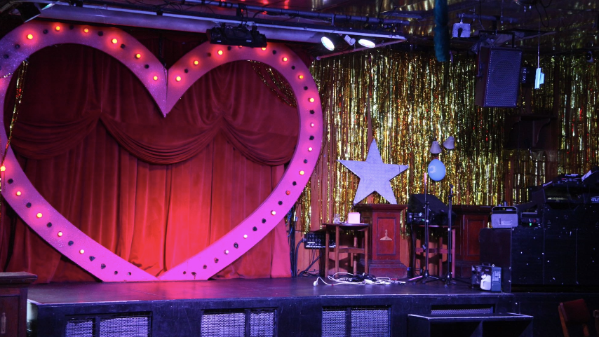 A stage with a large pink heart with gems around it against red curtains, next to gold foil curtains with a white star propped up against the wall.  