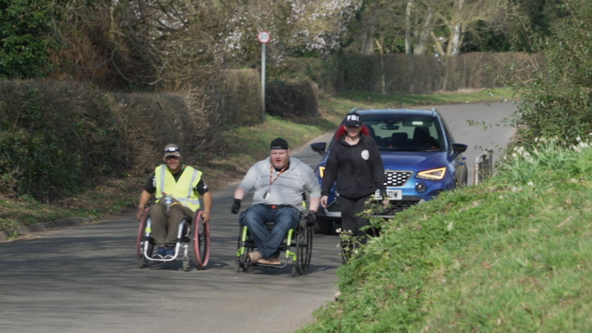 A woman in black clothing and a baseball cap walking on a country lane beside two men in wheelchairs, one in hi-vis and dark clothing, the other in a grey hoodie and dark trousers. All three are wearing baseball caps. A blue car is following them