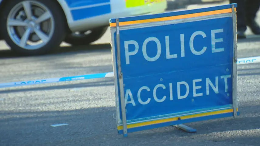 A blue sign with the words "Police Accident" in white writing. The sign is a rectangle, propped up on a blue stand, with a yellow border at the top and bottom. It is placed on the road in front of blue and white police tape. In the background, the wheel of a police car, with blue, white and luminous green trim of the car and the feet of a person wearing dark-coloured clothing visible.