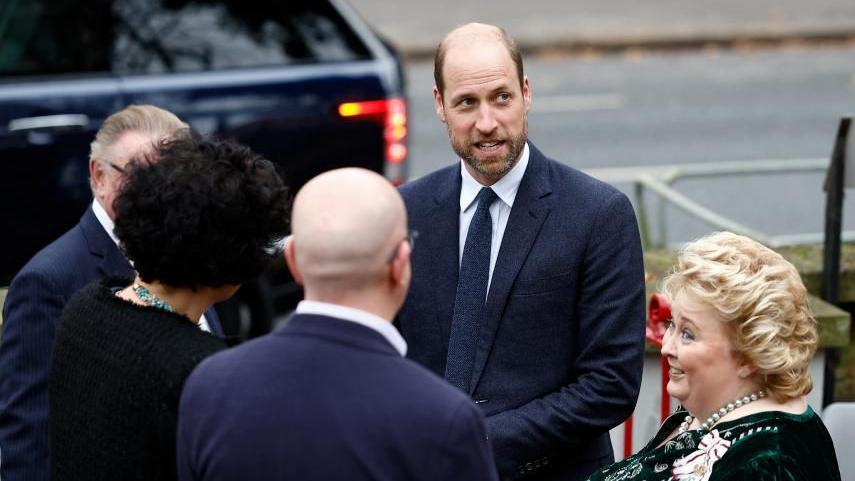 Prince William is standing with four other people who are not facing the camera. The Prince is smiling and wearing a dark coloured suit. 