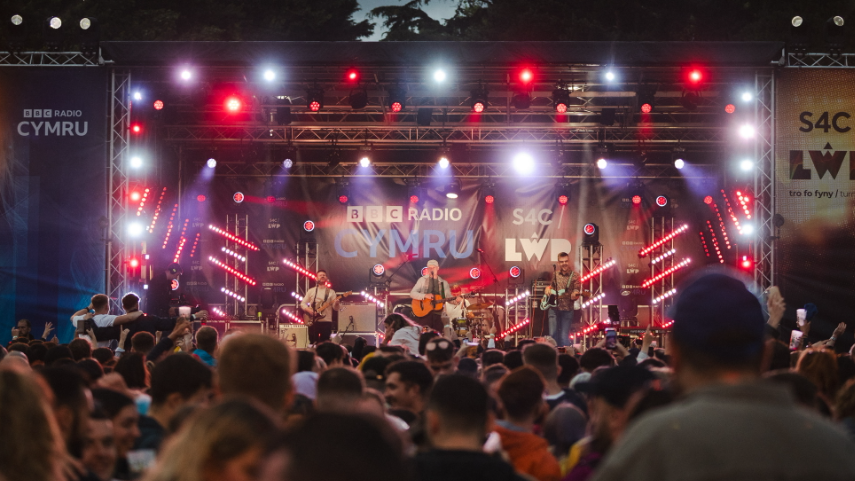 A stage with pink and purple lights at nightime, with a band on the stage including a guitarist in the middle and then the backs of the heads of the vast crowds in the foreground