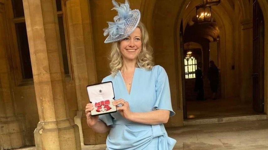 A picture of a woman wearing a sky blue formal dress and matching shoes. She has blonde hair and is holding her MBE award up to the camera.