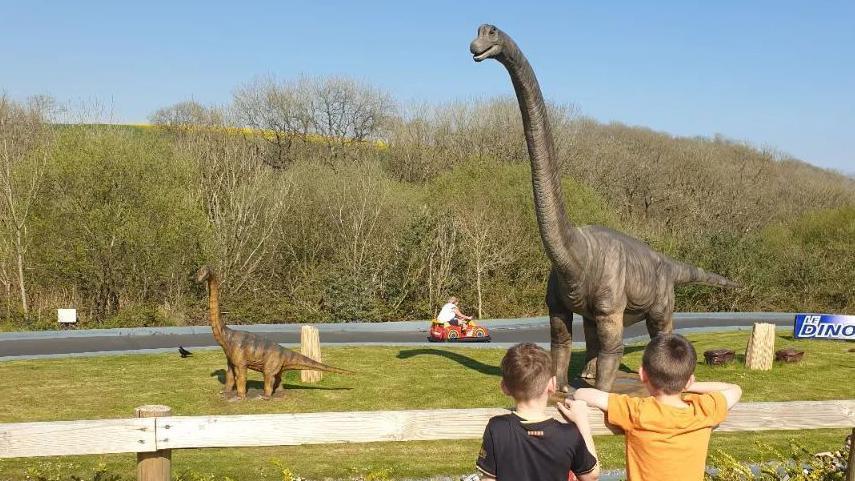 Two children stare at a giant model dinosaur while a go-cart goes by the background.