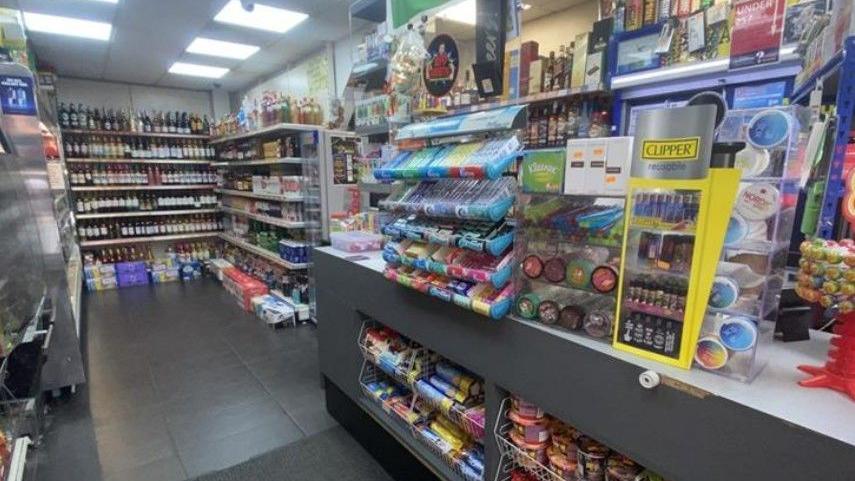 The shop counter is filled with products for sale including chewing gum and biscuits. Shelves further back are filled with products including bottles of wine.