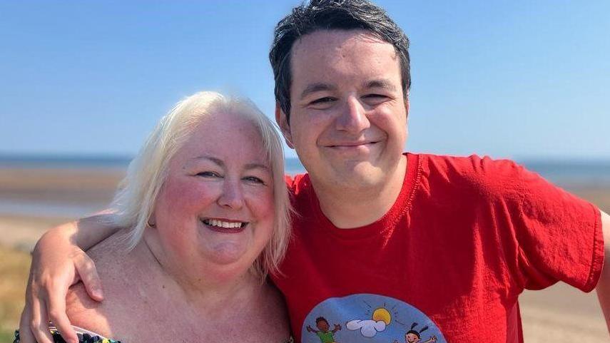 Beth, with bright blonde hair, smiles at the camera while her son, Calum, hugs her on a beach 