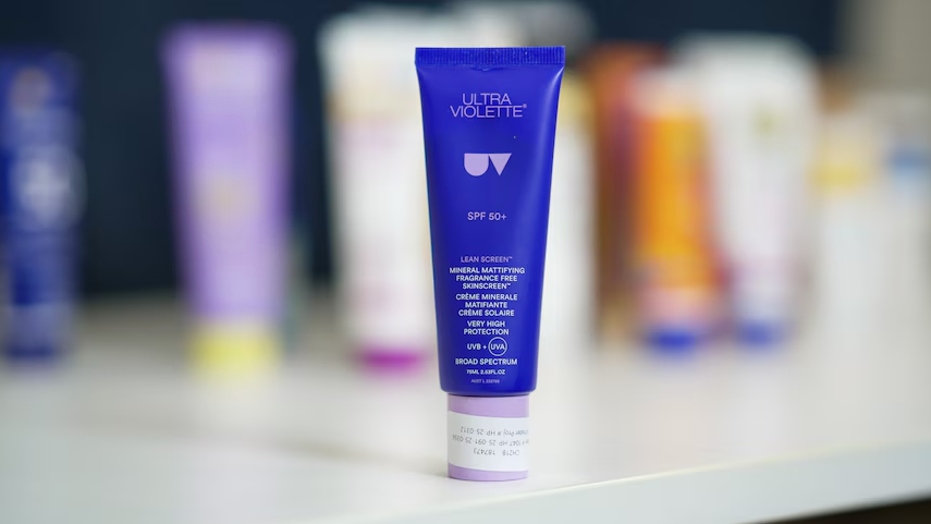 A purple and blue tube of sunscreen stands on a counter, with a range of other sunscreens blurred in the background