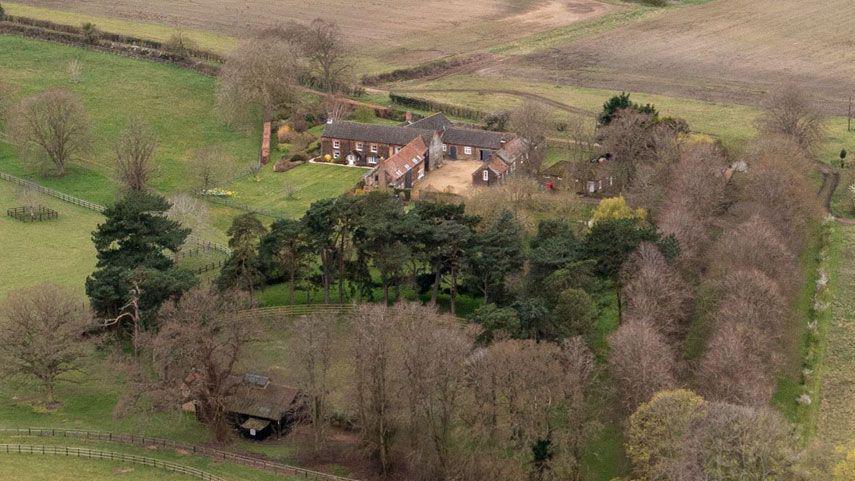 An aerial view of Wood Farm on the Sandringham estate. It is a large farmhouse hidden behind rows of trees and situated among roling fields.