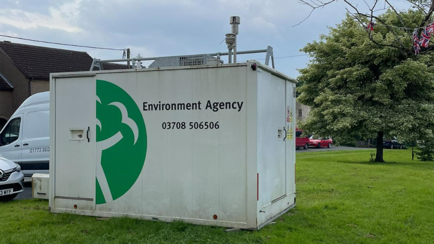 A large white unit with the words 'Environment Agency' written in black letters, situated on grass with a tree in the background.