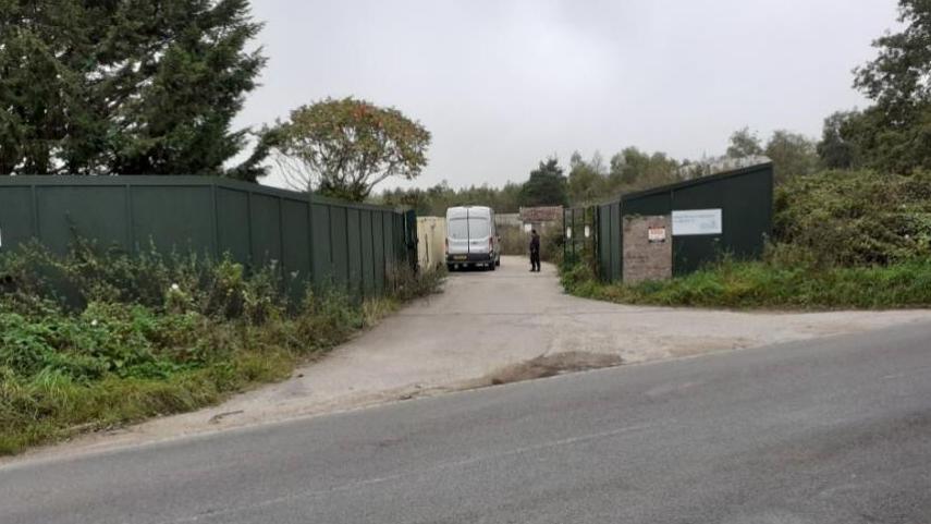 Dark green hoarding is either side of the entrance which is approached on a concrete driveway. A white van can be seen just inside the entrance and a person wearing dark clothing standing near it inside the entrance gates