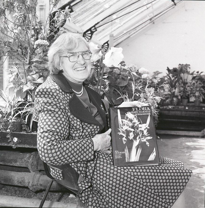 A black and white image of Ms Crackles holding her book, she is seated in a greenhouse and is wearing a skirt and jacket in spotted fabric.