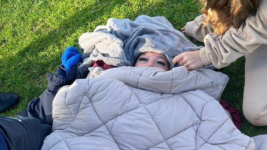 A young woman whose face is mostly obscured by warm coats and blankets, lies on grass while being attended to by people on their knees