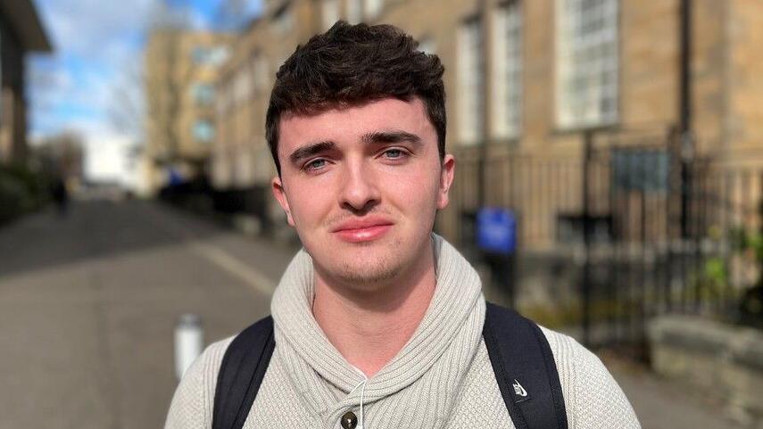 A man with short dark hair smiles at the camera. He wears a white jumper and a dark backpack. He is outside on the street and it is bright and sunny. 