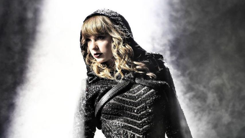 Taylor Swift wearing black glittered costume performs on stage.