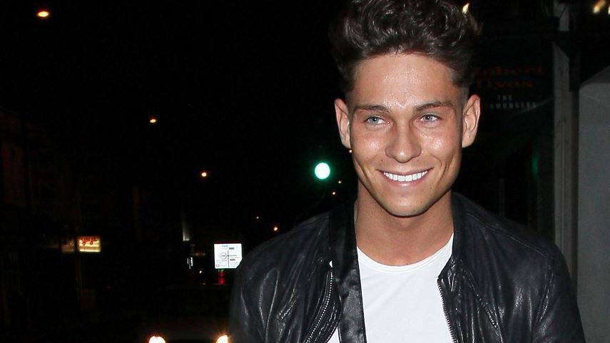 Joey Essex standing outside Sugar Hut at night. He has brown hair brushed backwards and is wearing a black leather jacket, unzipped to show a white T-shirt.