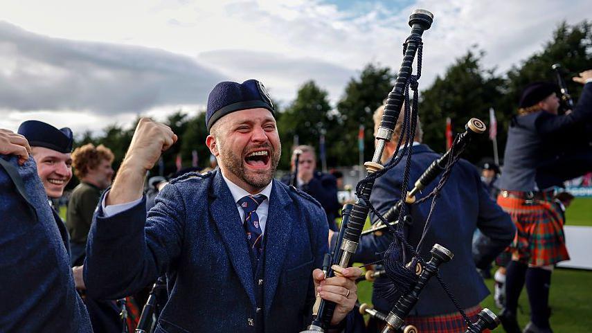 A piper from the Simon Fraser University pipe band celebrating their win. His mouth is open in a wide smile and he is punching the air. He is wearing a traditional bagpiper outfit with hat and is holding his bagpipes. There are other pipers behind him.