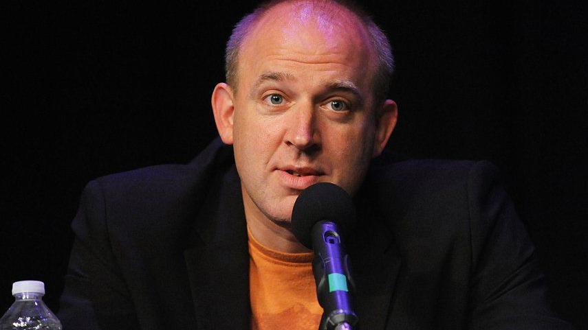 Comedian Tim Vine. He is wearning a orange t-shirt with a black jacket over the top. He is speaking into a microphone