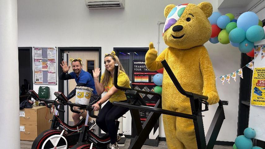Two people on an exercise bike wave at the camera and wear yellow bear ears. Next to them on a treadmill is Pudsey Bear, a giant yellow bear with an eye patch. He is doing a thumbs up.