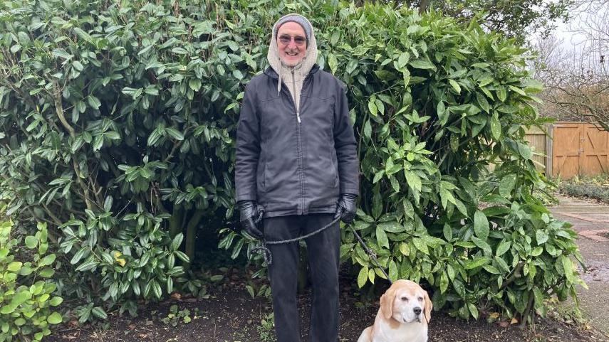 A man is standing in front of a large piece of shrubbery with a black jacket and trousers and a beige hoodie. He is holding a beagle on the lead and smiling at the camera.