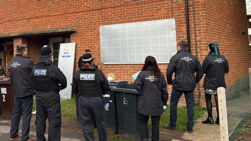 Police officers and council staff stand in front of a house with a silver steel place secured over the window.