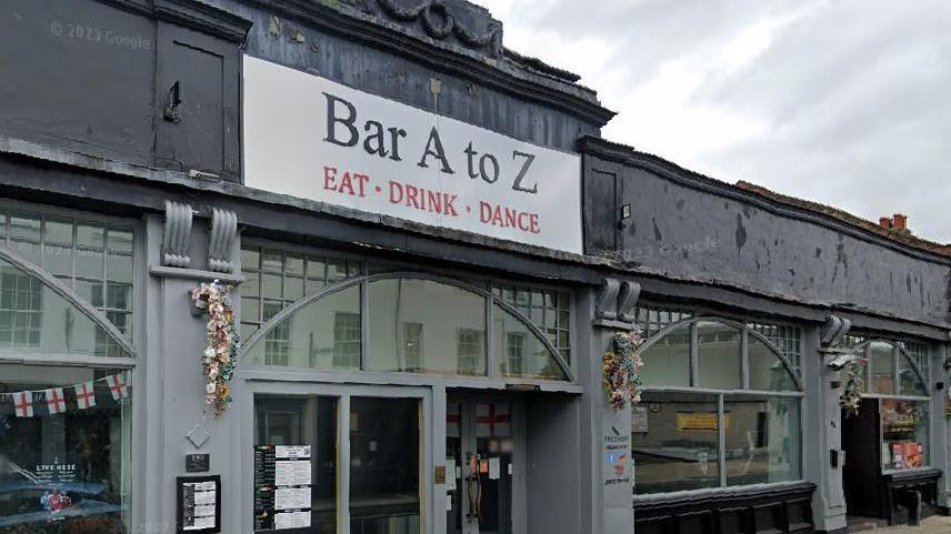 Bar A to Z is a black and grey building with large windows spanning the width of the building. The name sign of the bar says "eat, drink, dance" in a red font underneath. 