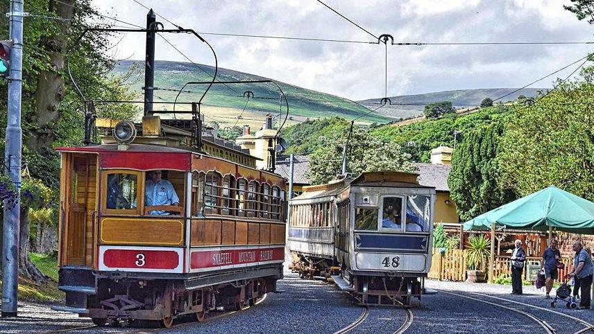 Two electric trams sit side by side on tracks with a canopy on the right and people waiting. In the background there are trees and rolling hills.