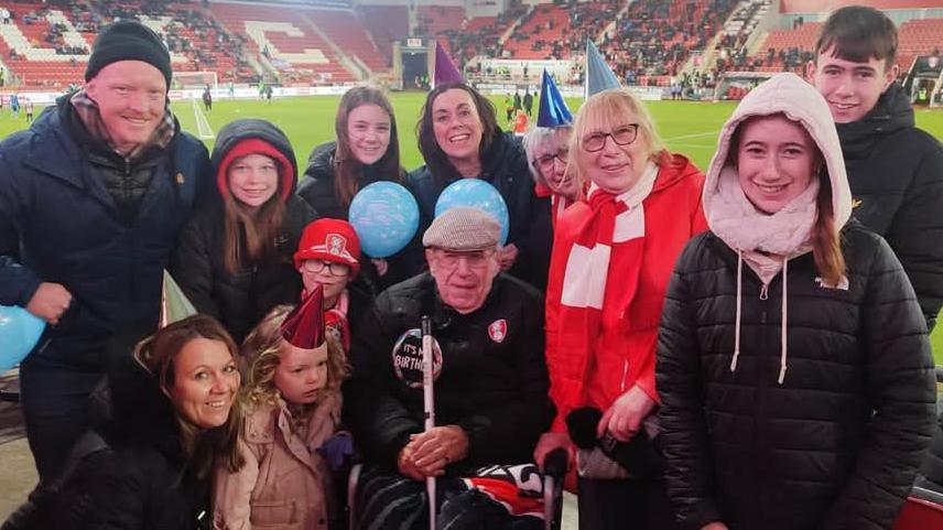 Dennis's family are gathered around him at Rotherham United's New York Stadium. He's celebrating his birthday, and wears a birthday badge on his chest. Several of his family are wearing party hats.