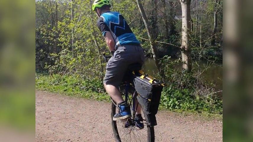 Luke Evison with his back to the camera, on his unicycle, cycling away from the camera.
He is wearing his black and yellow cycling helmet and a blue and black cycling t shirt and black long shorts with blue and black trainers. He is on a mud path with trees to the side.
