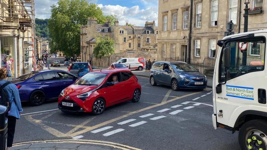 The busy Gay Street junction in Bath with queues of cars, including a red, blue and black car. There is also a Bath and North East Somerset lorry waiting at a junction, with cream-coloured homes in the background.