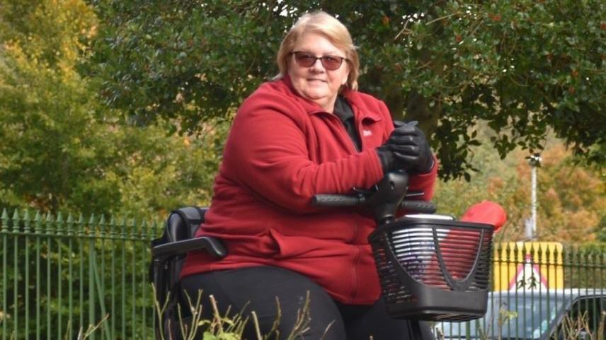 A woman in a red fleece coat is sitting on a mobility scooter.  She has shoulder length, light brown hair, and is wearing sunglasses.  She is on a path in the middle of a park and is surrounded by trees and greenery.