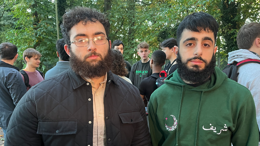 Noordin Jamal on the left and Sharief Younis stood to his right. Both men are looking at the camera with solemn expressions and behind them is a crowd of other students engaged in a conversation. Noordin Jamal wears a navy jacket with a beige shirt underneath and has thin rectangle framed glasses. Sharief Younis is wearing a dark green hoodie.