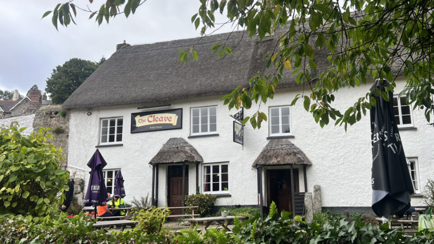 Outside The Cleave Inn in Lustleigh. The property is white with a thatched roof. Benches are outside the pub.