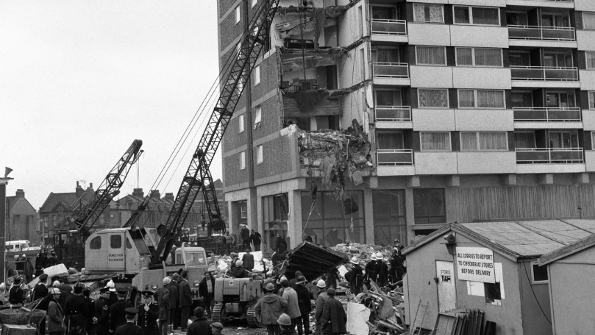 A black and white photo showing  multiple collapsed floors and walls on the corner of Ronan Point, with cranes stretching into the sky on the left. Crowds of people stand in front of a large pile rubble on the ground