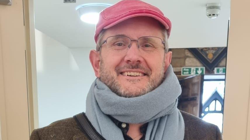 A smiling man with a beard and glasses wearing a red flat cap and light  blue scarf