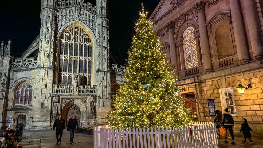 A large, well-lit Christmas tree is standing in front of Bath Abbey under the nightsky. There are people walking around it