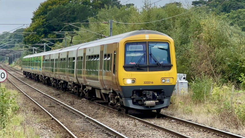 A yellow train with two carriages on track running through a wooded area with overhead wires overhead