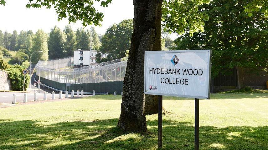 A sign for Hydebank Wood College in front of trees with the wire fencing surrounding the facility in the background