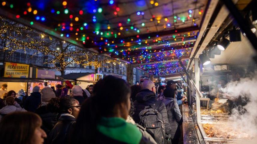 Multi-coloured fairy lights twinkle above festive stalls selling food and drink, with crowds of people walking past and waiting to be served.