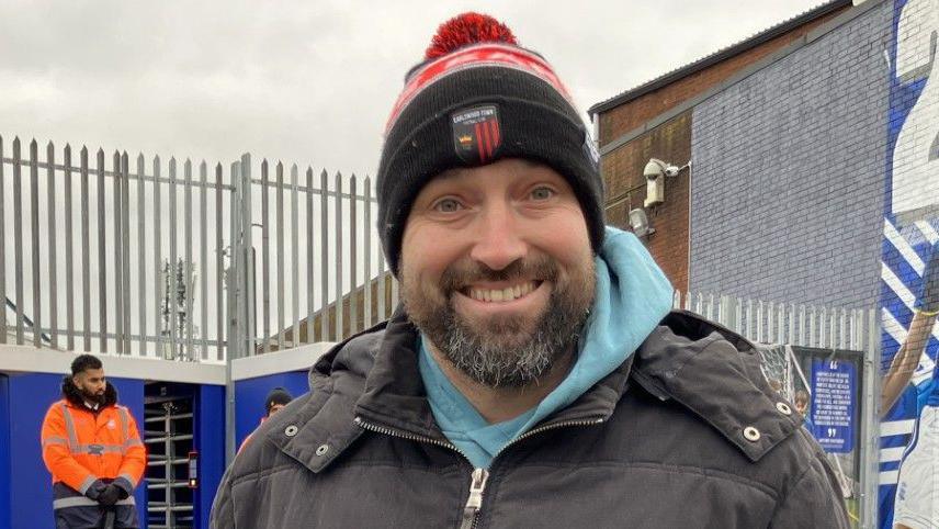 James Bowater smiling and wearing a beanie hat. He has a coat and a blue hoodie on. There is a steward behind him. 