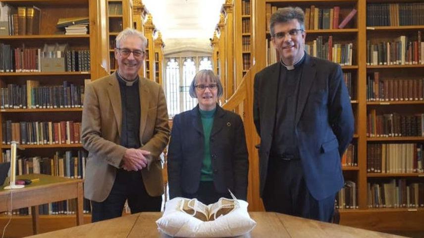 Three people - two men and a woman - stand at a table on which is an ancient bible in protective wrapping. All three are wearing clerical collars and smart outfits and are smiling at the camera