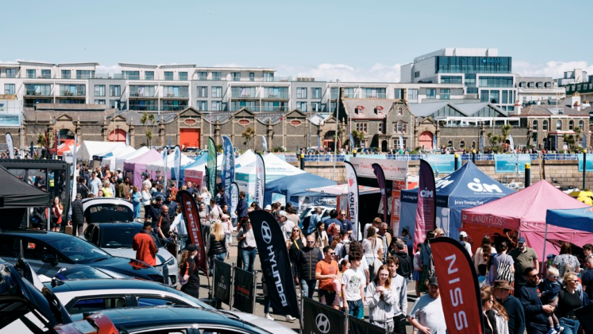 High angle photo of a bustling pier at Jersey Boat Show. Lots of people are walking up and down a sunny promenade as pop up business tents and flags line the periphery