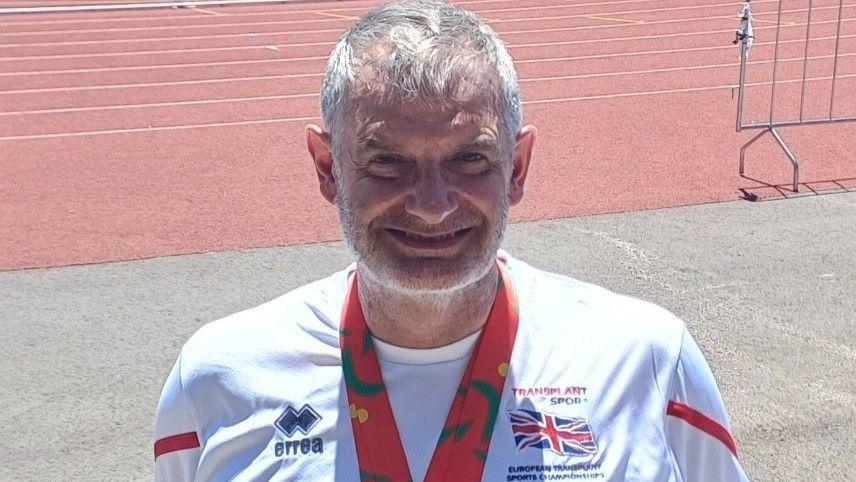 Rob Hodgkiss wearing his medals around his neck in front of the running 