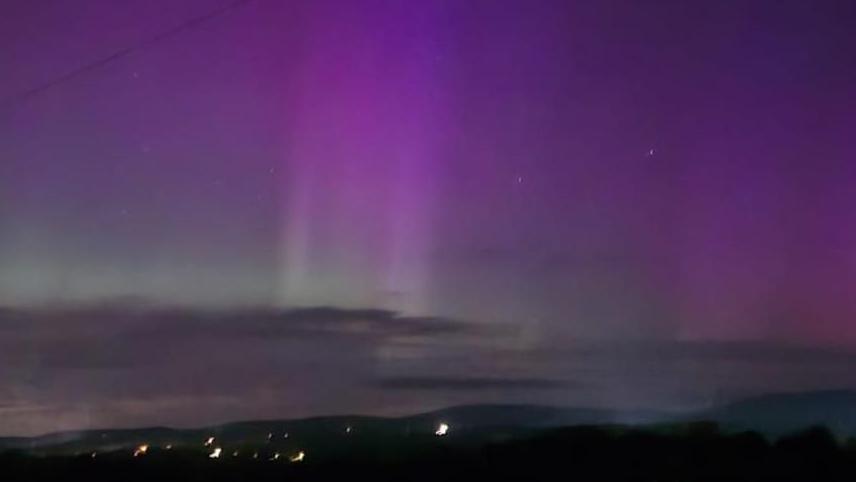 Two purple beams of light can be seen in the night sky with stars and clouds. Below  there are scattered lights, grass, mountains and a silver gate.