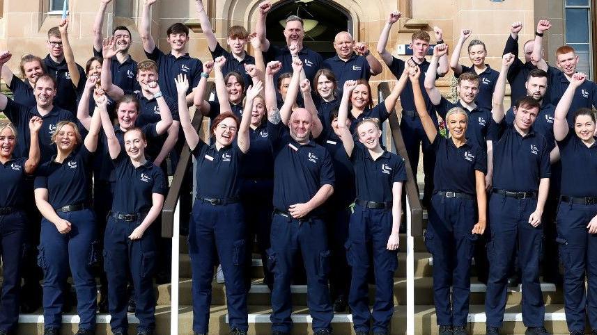 Shows young paramedics wearing blue trouser and t-shirts punching the air at the front of the main steps of Magee