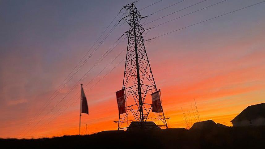 A metal electricity pylon with wires coming from its top is highlighted by the red, yellow and orange stripes in the sky.