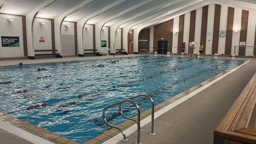 An indoor swimming pool with people swimming in the lanes, which are separated by red dividers. The back wall has white and brown stripes