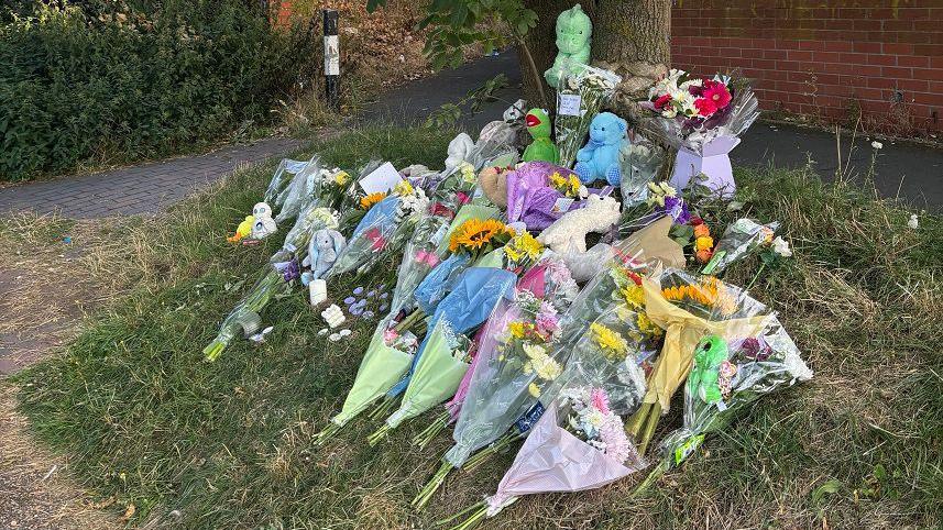 Bouquets of flowers lie on an area of grass next to a tree with a red brick wall visible in the background