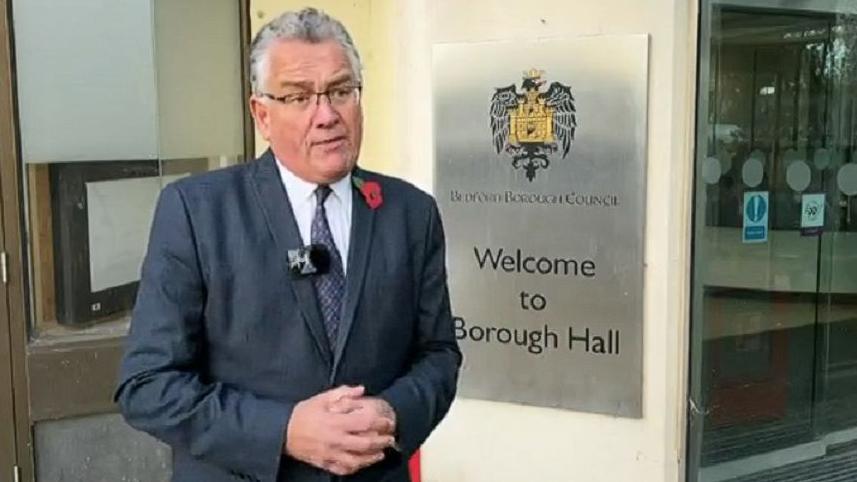Doug McMurdo with short grey hair and black-framed glasses wearing a grey jacket, white shirt and blue tie. He is wearing a red poppy and is standing in front of the entrance to Borough Hall. A sign behind him says "Welcome to Borough Hall".