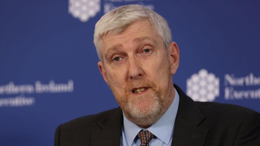 John O'Dowd has short grey hair and a grey/ginger beard. He is wearing a dark suit jacket. blue shirt and patterned tie
